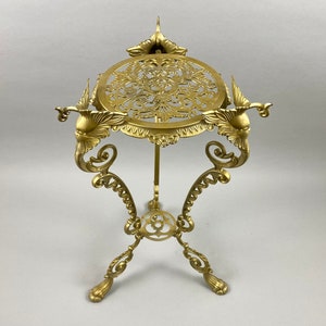 May include: A gold metal decorative stand with a round top and three legs. The stand features intricate floral and bird designs.