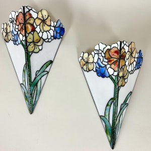 May include: Two stained glass wall sconces in a triangular shape, each with a floral design. The flowers are in orange, yellow, and blue, with green stems and leaves. The sconces are mounted on a light-coloured wall.