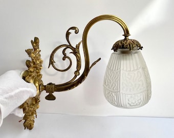 Può includere: Un'applique da parete in ottone con un paralume in vetro smerigliato. L'applique ha un design floreale e un braccio curvo. Il paralume ha un motivo geometrico.
