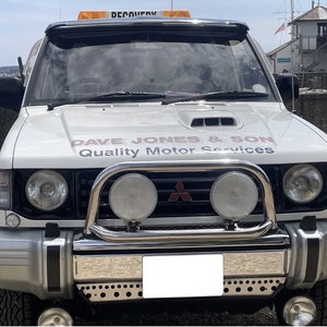 May include: White Mitsubishi Montero SUV with a silver front bumper, black grille, and two round headlights. The vehicle has a black bull bar with two additional round headlights. The vehicle has a sign on the bonnet that reads "Dave Jones & Son Quality Motor Services".