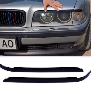 Pode incluir: Um par de peças de acabamento de sobrancelha de farol de plástico preto para um carro. As peças de acabamento são curvas e têm um acabamento brilhante.