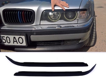 Könnte beinhalten: Ein Paar schwarze Kunststoff-Augenbrauenverkleidungen für einen Scheinwerfer eines Autos. Die Verkleidungen sind gebogen und haben eine glänzende Oberfläche.
