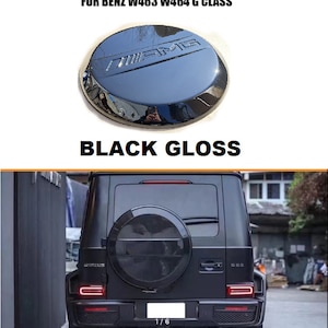 May include: Black gloss spare tire cover with the Mercedes-Benz AMG logo for a black Mercedes-Benz G-Wagon. The cover is mounted on the back of the car.