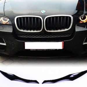 Puede incluir: Un par de cubiertas de faros delanteros de plástico negro para un BMW X6. Las cubiertas están diseñadas para proteger los faros de los daños y para darle al coche un aspecto más agresivo.