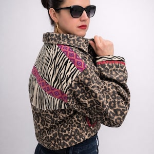 May include: A cropped jacket with a leopard print pattern, zebra print, and fringe detailing. The jacket features a collar and long sleeves with a red and black trim. The model is wearing black sunglasses and dark blue jeans.