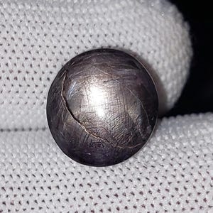 Op de afbeelding: Een donkere, ronde edelsteen met een gebarsten oppervlak, tegen een witte stoffen achtergrond. De edelsteen heeft een metallic glans en lijkt een gepolijste steen te zijn. De afbeelding is goed verlicht en benadrukt de details van de edelsteen.