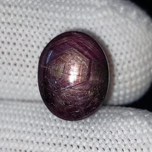 May include: A dark purple, oval gemstone with a star effect. The gem is set against a white fabric background. The stone has a polished surface with visible internal structures, reflecting light.