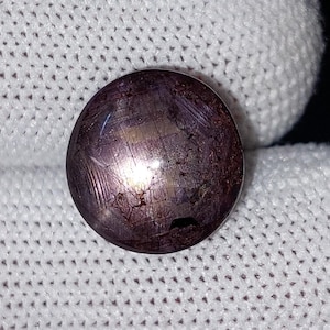 Op de afbeelding: Een ronde, gepolijste edelsteen met een dieppaarse tint, die een stervormig effect over het oppervlak vertoont. De edelsteen wordt tegen een witte stoffen achtergrond gehouden, wat de kleur en textuur benadrukt.