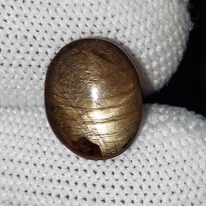 Op de afbeelding: Ovale, goudbruine edelsteen met een gepolijst oppervlak. De steen heeft een kenmerkend, wervelend patroon en een kleine, donkere insluitsel. De edelsteen wordt tegen een witte, getextureerde achtergrond gehouden.