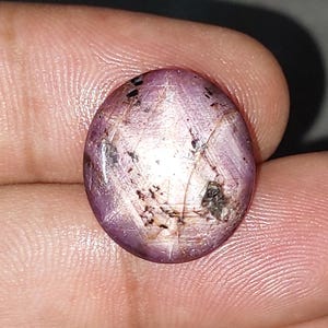 May include: Oval-shaped, purplish-pink gemstone with a star-like pattern. The stone has a polished surface with dark inclusions and a subtle sheen. The gem is held between fingers, showcasing its color and texture. A unique piece for jewelry.