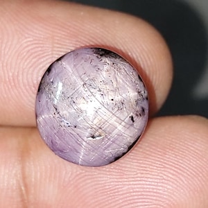 May include: A round, cabochon-cut star sapphire gemstone with a translucent, pale lavender-purple hue. The stone features a visible six-rayed star effect across its polished, domed surface, along with natural internal inclusions and dark mineral flecks.