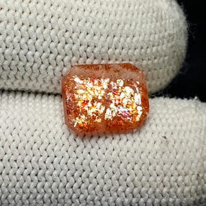 Peut inclure: Une pierre précieuse de type pierre de soleil de forme rectangulaire, à la surface polie. La pierre présente une teinte brun-orangé chaud avec des inclusions chatoyantes et réfléchissantes. La pierre est posée sur un fond blanc tricoté.