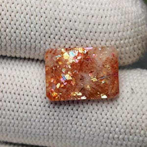 Könnte beinhalten: Ein rechteckiger Sonnenstein-Edelstein mit einer warmen orange-braunen Grundfarbe und eingebetteten metallischen, irisierenden Flocken, die in Gold-, Rosa- und Blautönen schimmern. Der Stein hat eine polierte, facettierte Oberfläche mit Einschlüssen.