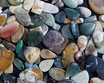 May include: A collection of polished gemstones in various shapes and colors. The stones include shades of brown, gray, green, pink, and black. Some stones have unique patterns and textures, with a mix of oval, teardrop, and irregular shapes.