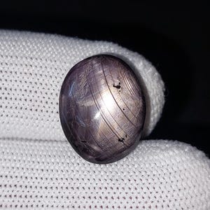 May include: A dark purple, oval gemstone with a polished surface. The stone displays light streaks and small inclusions. The gem is set against a white background, accentuating its colour and texture. A close-up shot.