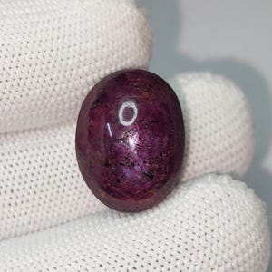 Op de afbeelding: Een gepolijste, ovaalvormige edelsteen met een diepe bordeauxrode kleur en een reflecterend oppervlak. De steen heeft donkere insluitsels en wordt tegen een witte achtergrond gehouden. Waarschijnlijk een robijn, geschikt voor sieraden.