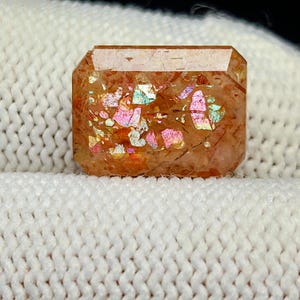 Op de afbeelding: Een rechthoekige edelsteen met een warme, oranje-bruine tint. De edelsteen heeft interne iriserende insluitsels die roze, blauw en geel licht reflecteren. De steen ligt op een witte, getextureerde achtergrond.