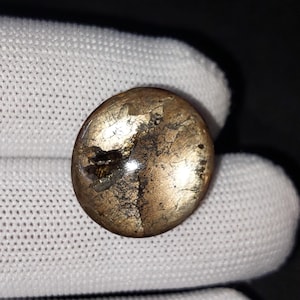 Op de afbeelding: Een ronde knoop in antieke stijl met een metallic gouden afwerking en donkere, getextureerde accenten. De knoop heeft een licht gewelfde vorm en een versleten uiterlijk, wat duidt op leeftijd en gebruik. De knoop is waarschijnlijk van metaal.