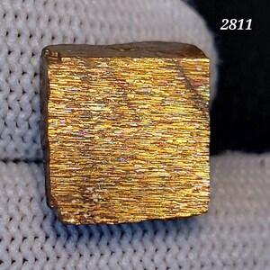 May include: A golden, cube-shaped mineral specimen with a textured, iridescent surface. The surface displays a pattern of horizontal lines and a range of colours, including gold, bronze, and hints of other hues. The number "2811" is visible in the upper right corner.