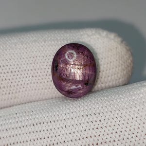 May include: A polished, oval gemstone in a deep pinkish-purple colour. The stone displays a star-like effect, with lines radiating from a central point. The gem is set against a white, textured background, highlighting its colour and unique internal structure.