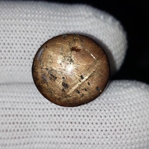 Op de afbeelding: Een ronde, antieke bronzen knoop met een versleten, getextureerd oppervlak. De knoop heeft een licht gewelfde vorm en vertoont tekenen van ouderdom, met krassen en donkere vlekken. Waarschijnlijk afkomstig van een vintage kledingstuk of collectie.