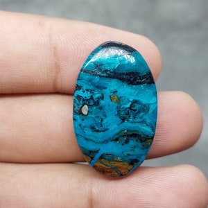 Może przedstawiać: Owalny kamień szlachetny o uderzającym wzorze w odcieniach turkusu, czerni i brązu. Wypolerowana powierzchnia ukazuje wirujący, warstwowy wzór z akcentami złota i małą, srebrną inkluzją. Kamień trzymany jest w dłoni.