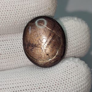 Op de afbeelding: Een ronde, metaalachtige kraal met een bronzen tint. Het oppervlak heeft een textuur met zichtbare krassen en onvolkomenheden. Een witte ring is zichtbaar op de bovenkant van de kraal. De kraal wordt vastgehouden door een witte gehandschoende hand.