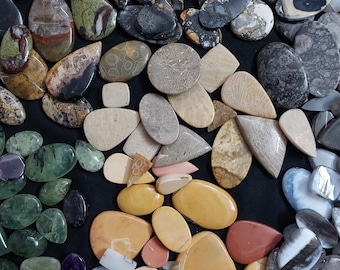May include: A collection of polished gemstones in various shapes and colors, including green, brown, yellow, and gray. The stones are arranged on a black surface, showcasing their unique patterns and textures. The assortment includes oval, teardrop, and irregular shapes.