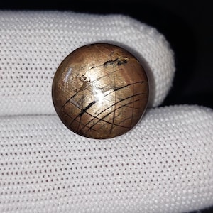 Op de afbeelding: Een ronde, bronskleurige knoop met een bekraste oppervlakte en zwarte lijnen. De knoop is geplaatst tegen een witte, getextureerde achtergrond. Het ontwerp van de knoop heeft een reeks kruisende lijnen, waardoor een uniek patroon ontstaat.