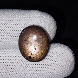 Op de afbeelding: Een ronde, gepolijste edelsteen met een metallic bruine kleur en een enigszins ongelijkmatig oppervlak. De steen heeft gouden vlekken en donkere vlekken, wat wijst op insluitsels of natuurlijke variaties. De edelsteen ligt tegen een witte, getextureerde achtergrond.