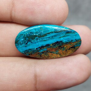 Op de afbeelding: Een ovaalvormige edelsteen met een opvallend kleurenpalet. De steen heeft een levendige turquoise basis, geaccentueerd met zwarte, bruine en gouden strepen. Het gepolijste oppervlak onthult een natuurlijk, getextureerd patroon, wat een unieke uitstraling creëert.