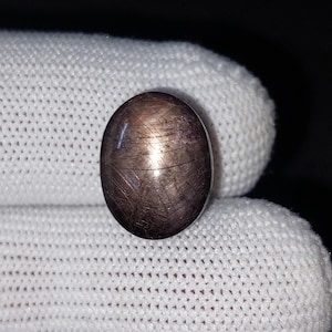 Op de afbeelding: Ovale, donkerbruine edelsteen met een ster-effect. Het gepolijste oppervlak reflecteert licht en toont een uniek patroon van kruisende lijnen. De edelsteen ligt op een witte stoffen achtergrond.