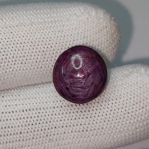 May include: A round, polished gemstone with a deep reddish-purple hue. The gem has a star-like pattern across its surface, and is set against a white background. The gemstone is likely a ruby, and is a beautiful example of natural mineral artistry.