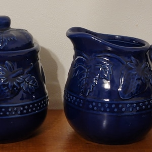 May include: A cobalt blue ceramic sugar bowl with a lid and a matching creamer pitcher. Both pieces feature embossed floral and leaf designs. The sugar bowl has a round shape, and the pitcher has a handle and spout.