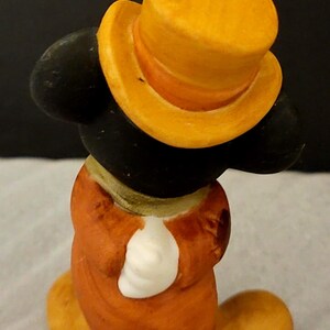 K&ouml;nnte beinhalten: Eine kleine Figur einer Zeichentrickmaus mit einem leuchtend gelben Zylinder, einem langen orangefarbenen Mantel und wei&szlig;en Handschuhen. Die Figur hat gro&szlig;e schwarze Ohren und ist von hinten dargestellt, wobei die R&uuml;ckseite von Mantel und Hut zu sehen ist.
