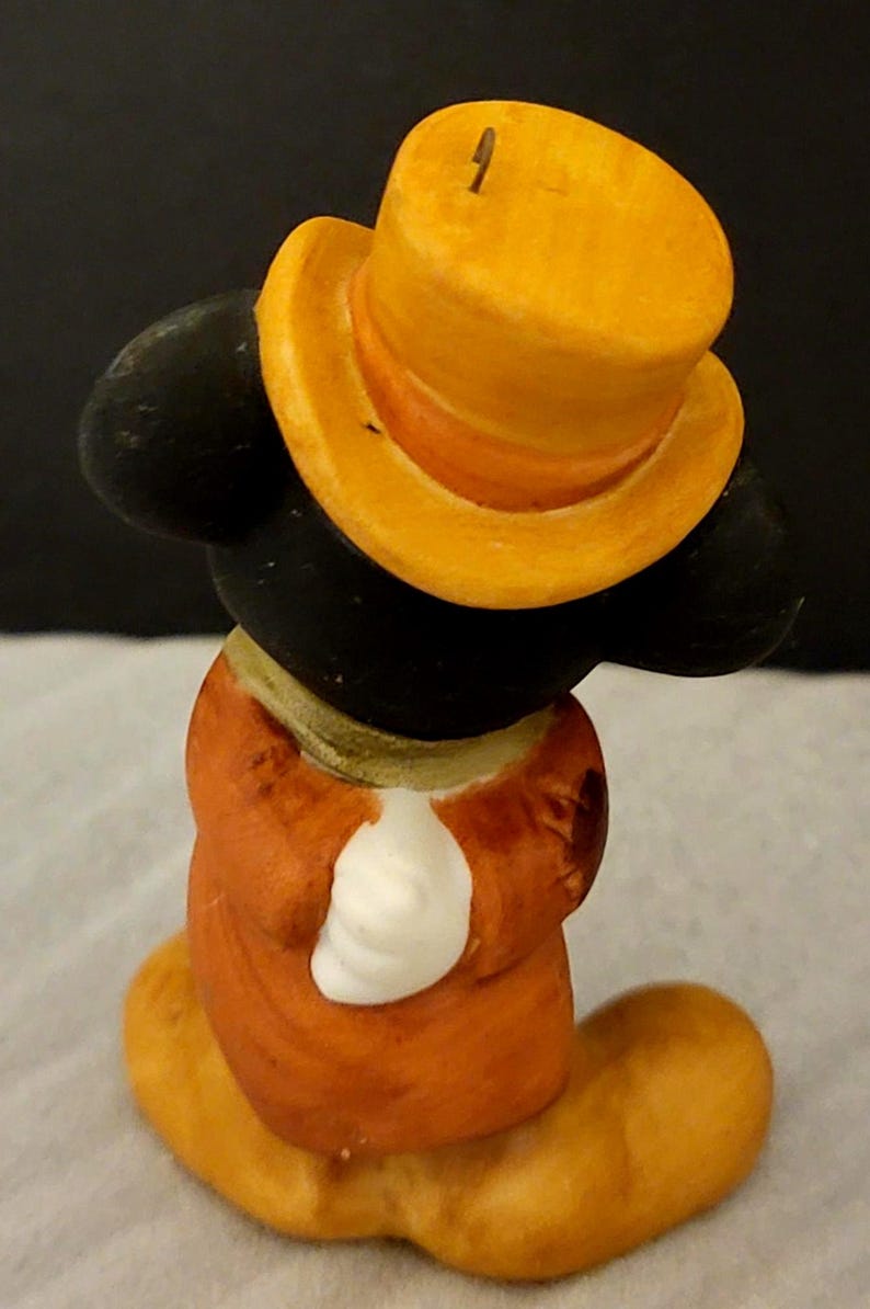 K&ouml;nnte beinhalten: Eine kleine Figur einer Zeichentrickmaus mit einem leuchtend gelben Zylinder, einem langen orangefarbenen Mantel und wei&szlig;en Handschuhen. Die Figur hat gro&szlig;e schwarze Ohren und ist von hinten dargestellt, wobei die R&uuml;ckseite von Mantel und Hut zu sehen ist.