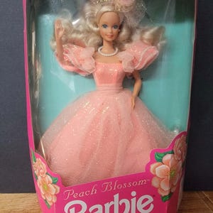 Puede incluir: Muñeca Barbie Peach Blossom en una caja rosa. La muñeca tiene cabello rubio, un collar de perlas y un vestido color melocotón con mangas con volantes. La caja presenta el logo de Barbie y detalles florales.