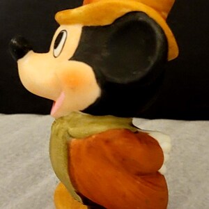 K&ouml;nnte beinhalten: Eine Keramikfigur von Mickey Mouse mit einem gelben Zylinder, einer gr&uuml;nen Weste und einer orangefarbenen Jacke. Die Figur ist im Profil dargestellt und zeigt einen schwarzen Kopf, ein wei&szlig;es Gesicht und gelbe Schuhe. Eine Metall&ouml;se ist am Hut befestigt.