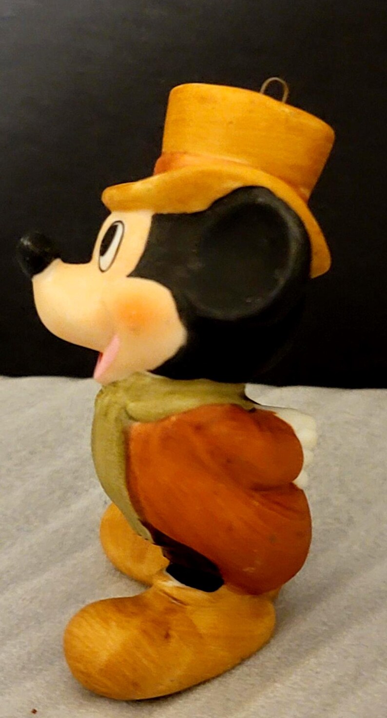 K&ouml;nnte beinhalten: Eine Keramikfigur von Mickey Mouse mit einem gelben Zylinder, einer gr&uuml;nen Weste und einer orangefarbenen Jacke. Die Figur ist im Profil dargestellt und zeigt einen schwarzen Kopf, ein wei&szlig;es Gesicht und gelbe Schuhe. Eine Metall&ouml;se ist am Hut befestigt.