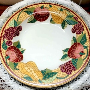 May include: A decorative ceramic plate with a white center and a mosaic-style border. The border features colorful illustrations of fruits, including apples, pears, and grapes, with green leaves. The plate has a yellow and tan background.