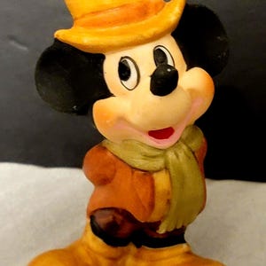 K&ouml;nnte beinhalten: Eine kleine Mickey-Mouse-Figur mit einem gelben Zylinder, einer braunen Jacke, einem hellgr&uuml;nen Schal und gelben Stiefeln. Die Figur hat schwarze Ohren, ein pfirsichfarbenes Gesicht und einen l&auml;chelnden Ausdruck mit einem sichtbaren roten Mund.