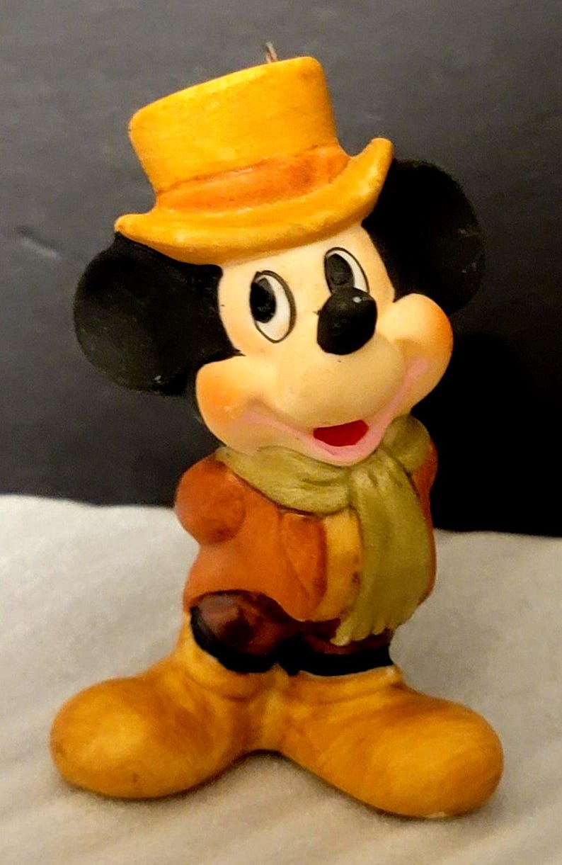 K&ouml;nnte beinhalten: Eine kleine Mickey-Mouse-Figur mit einem gelben Zylinder, einer braunen Jacke, einem hellgr&uuml;nen Schal und gelben Stiefeln. Die Figur hat schwarze Ohren, ein pfirsichfarbenes Gesicht und einen l&auml;chelnden Ausdruck mit einem sichtbaren roten Mund.