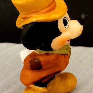 K&ouml;nnte beinhalten: Eine kleine Keramikfigur einer Zeichentrickmaus mit gelbem Zylinder, braunem Mantel und gelben Schuhen. Die Figur hat einen schwarzen Kopf, gro&szlig;e runde Ohren und eine cremefarbene Schnauze. An der Oberseite des Zylinders befindet sich eine Metall&ouml;se.
