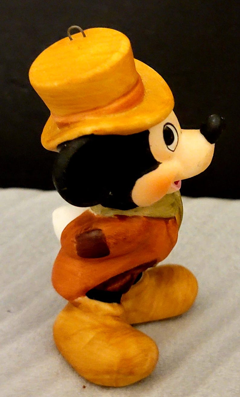 K&ouml;nnte beinhalten: Eine kleine Keramikfigur einer Zeichentrickmaus mit gelbem Zylinder, braunem Mantel und gelben Schuhen. Die Figur hat einen schwarzen Kopf, gro&szlig;e runde Ohren und eine cremefarbene Schnauze. An der Oberseite des Zylinders befindet sich eine Metall&ouml;se.