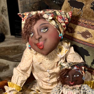 May include: Two handmade cloth dolls. The larger doll wears a yellow floral dress and headscarf. The smaller doll wears a blue floral dress and apron. Both dolls have dark skin, painted faces, and are seated.