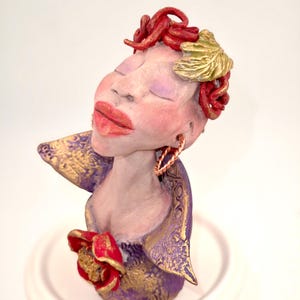 May include: A handcrafted ceramic sculpture of a woman with closed eyes, red hair, and gold accents. The sculpture features a red rose and is painted in shades of purple, gold, and red. The figure is mounted on a white base.