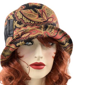 May include: A paisley print cloche hat in black, red, and gold. The hat features a small brim and a black accent on the side. The hat is displayed on a mannequin head with red hair and red lipstick.