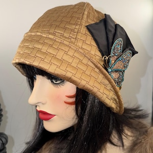 May include: A gold-colored cloche hat featuring a woven-look texture and a black fabric bow accent. The hat is adorned with a decorative butterfly brooch encrusted with small blue and bronze-toned crystals on the right side of the brim.