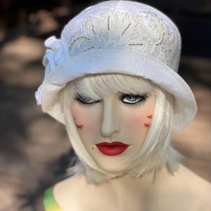 May include: A white cloche hat with a floral design and beaded accents. The hat features a small, white fabric flower on the side. The hat is on a mannequin head with a wig and makeup.