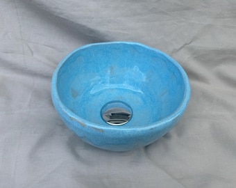 May include: A round, hand-formed ceramic vessel sink featuring a vibrant, glossy turquoise blue glaze with subtle tonal variations. The basin has an irregular, organic rim and includes a polished, reflective chrome-finished drain stopper at the center.